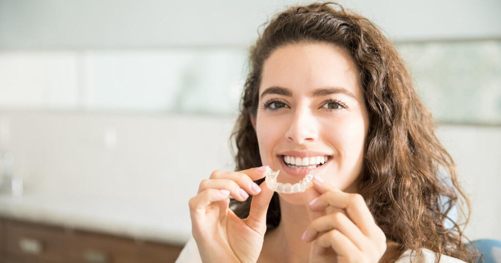 Smiling adult woman holding an Invisalign clear aligner in front of her mouth