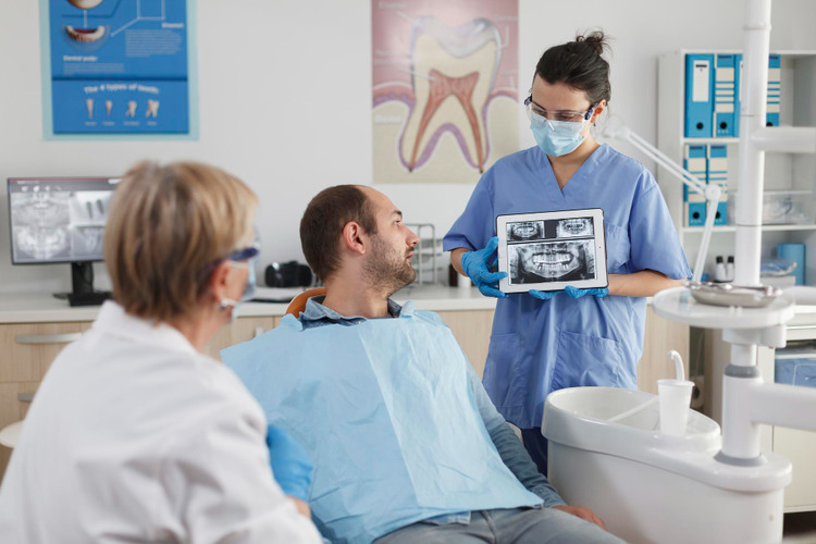 Dental clinic setting, male patient sitting on the dental chair, female dentist sitting beside him, and dental assistant on the other side holding a dental X-ray image