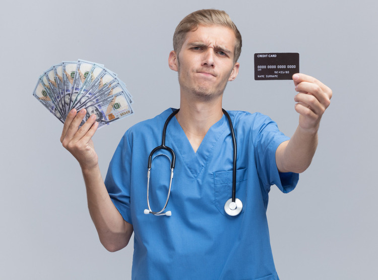 Male dentist holding cash and a bank card in his hand, with a worried and unhappy expression on his face.