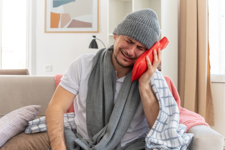 Man sitting on a sofa at home holding a compress on his cheek due to tooth pain from a delayed cracked tooth treatment.