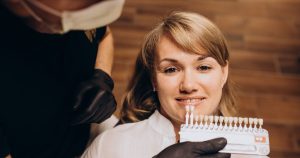 A woman is lying back on a dental chair, smiling, while a dentist is holding a set of porcelain veneers in front of her teeth.
