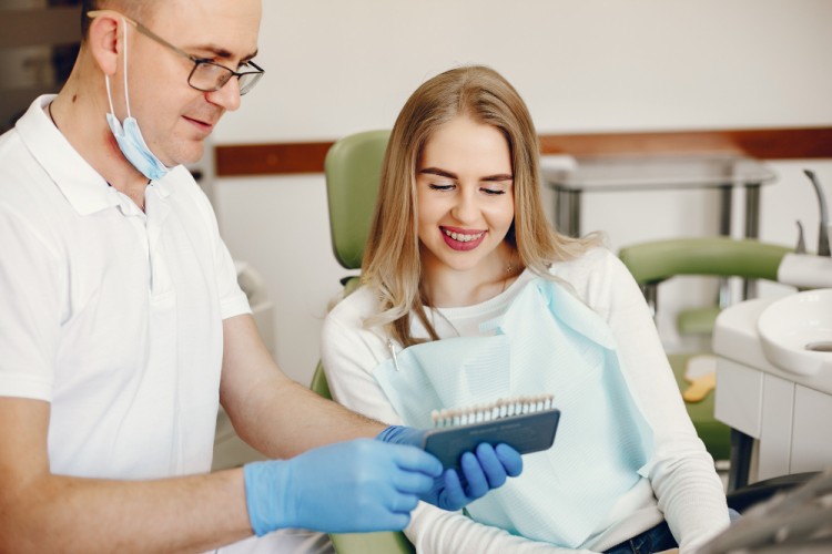 A male dentist in a clinic holding porcelain veneers in his hand while consulting and explaining the treatment to a female patient.