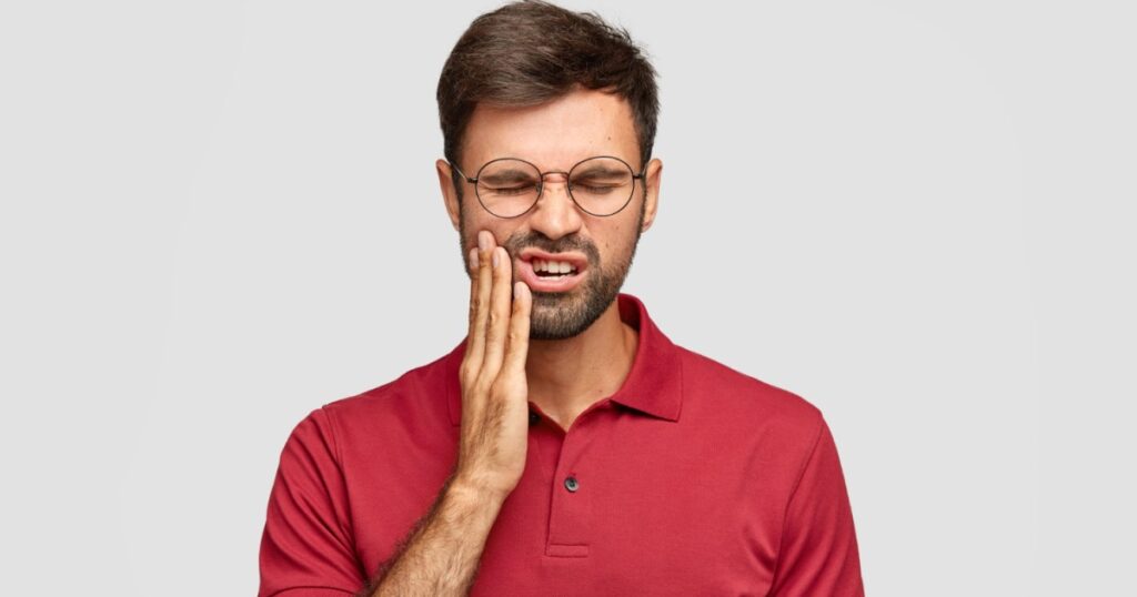 Young man wearing glasses experiencing a chipped tooth, holding his cheek in pain with eyes closed due to discomfort.