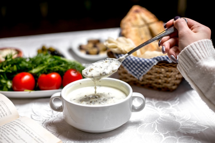 A bowl of soft white food on a table, with a hand gently scooping it with a spoon, while other foods are blurred in the background, suitable for a chipped tooth recovery diet.