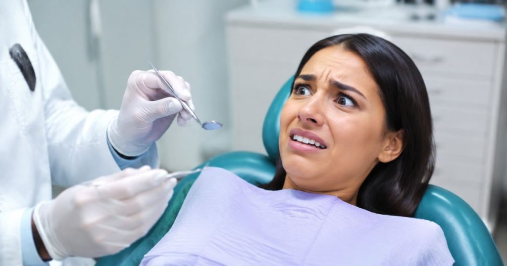 A young woman sitting on a dental chair showing signs of dental anxiety, with a frightened and anxious expression while looking at dental instruments in the dentist’s hands.