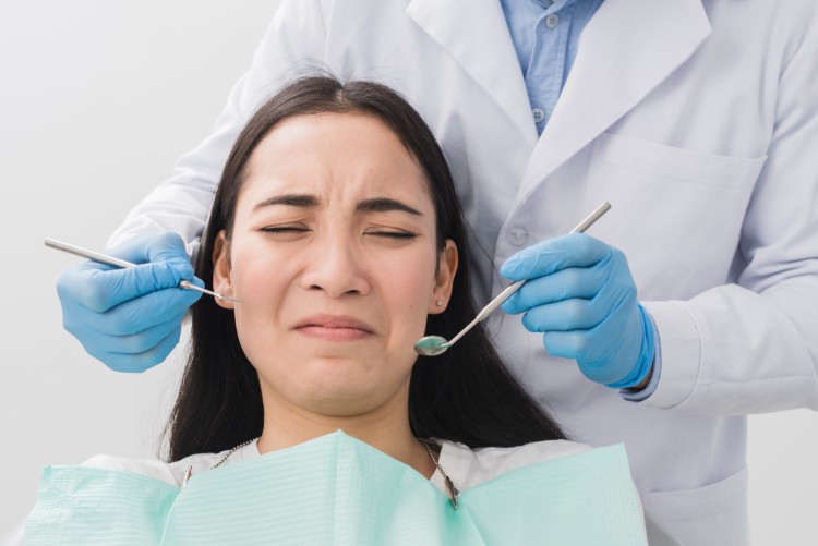 A young woman with her eyes tightly closed due to severe dental anxiety and fear, while a dentist holds dental instruments in front of the patient’s face.