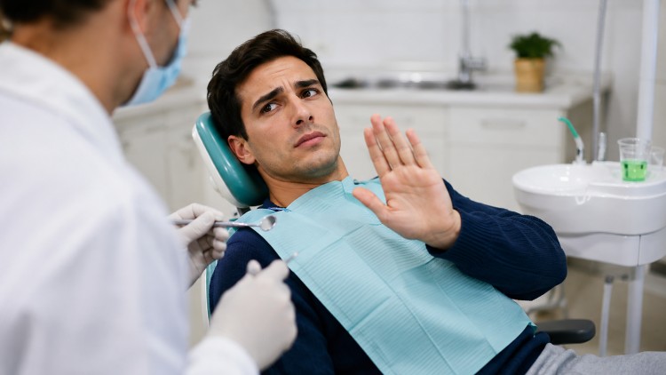 A young man undergoing dental treatment, raising his hand in distress due to dental anxiety and signaling “stop” to the dentist with his open palm.