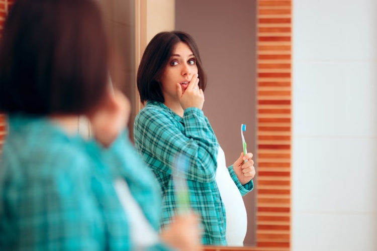 Pregnant woman brushing her teeth while looking in the mirror, with a hand on her mouth, focusing on oral hygiene and gum health during pregnancy.