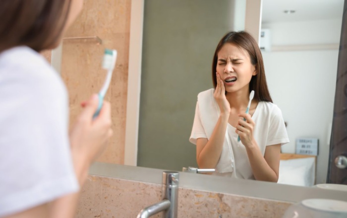 A young woman unhappy with her existing dental bridge, experiencing pain while brushing her teeth in front of the bathroom mirror.